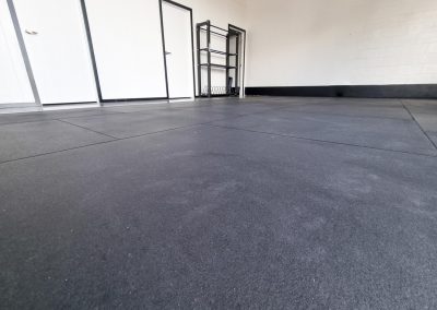 The empty room features Gym Tiles with black rubber flooring, pristine white walls, and a sturdy metal rack against the wall. Two closed white doors complete this sleek setup ideal for fitness spaces in Auckland